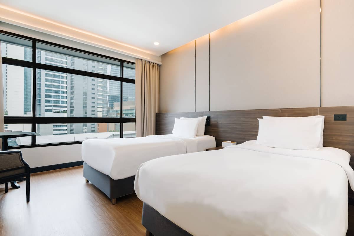 A hotel room with two beds, a desk, and a large window with a city view.