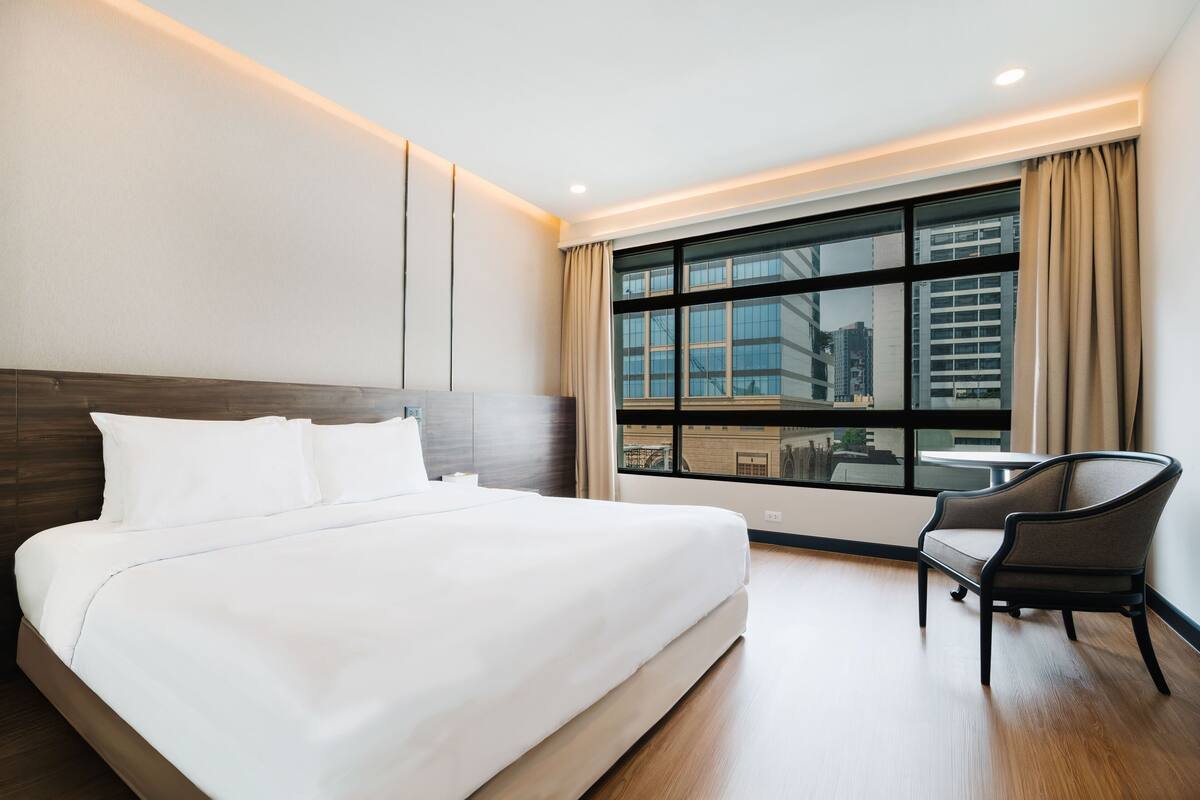 A hotel room with a large bed, a chair, a window with a city view, and a modern design.