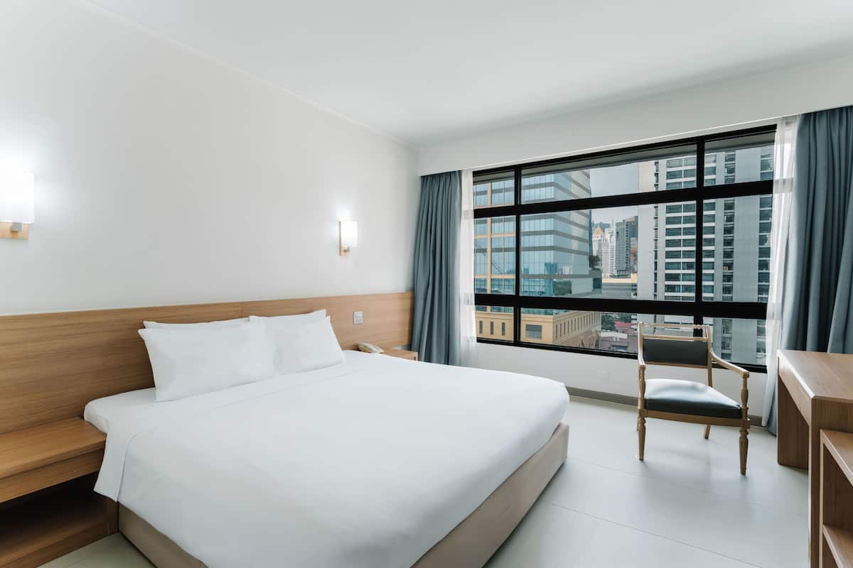 A hotel room with a large bed, a wooden nightstand, a desk, a chair, and a large window with a city view.