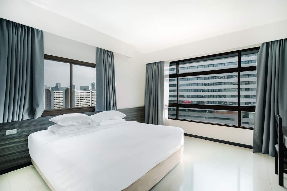 A modern hotel room with a large bed, two windows with curtains, and a city view.