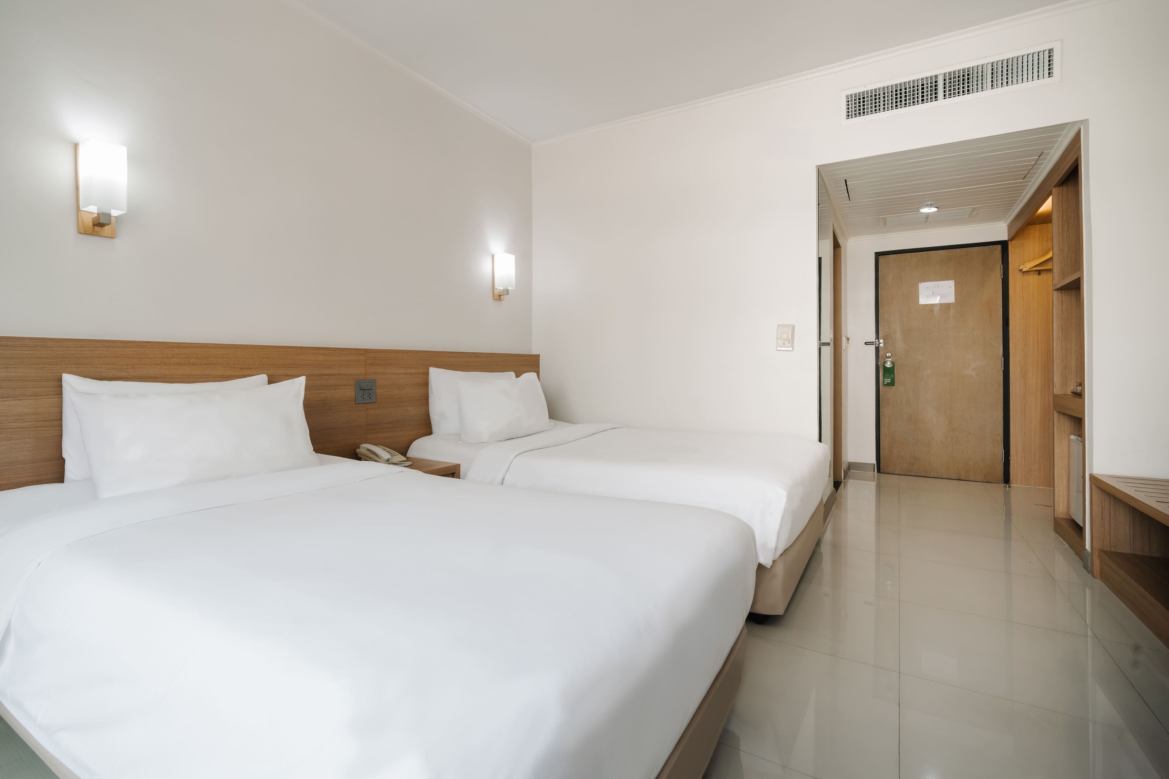 A hotel room with two beds, a wooden headboard, a bedside table with a phone, and a door leading to another room.