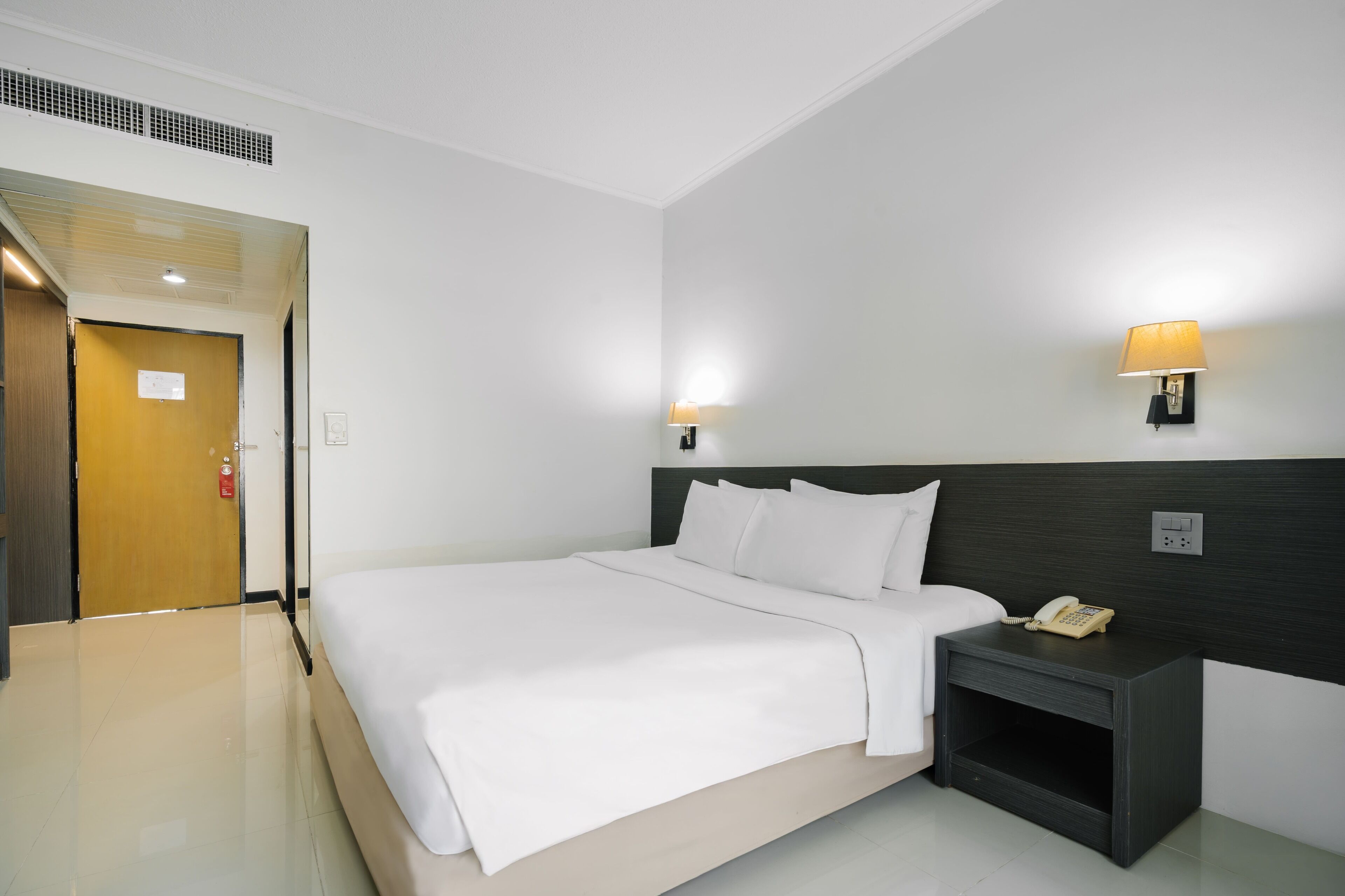A hotel room with a bed, bedside table with a phone, two wall-mounted lamps, a door, and a vent.