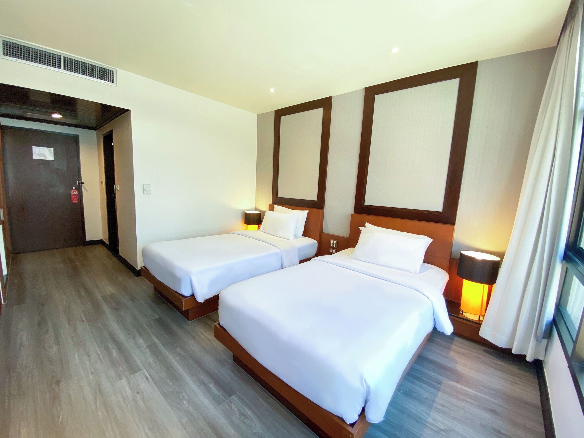 A hotel room with two single beds, a wooden floor, a large window with curtains, and a door with a fire extinguisher.