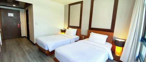 A hotel room with two single beds, a wooden floor, a large window with curtains, and a door with a fire extinguisher.