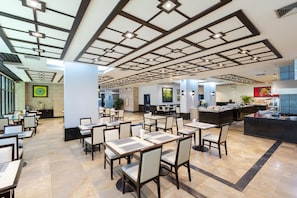 Un vaste espace dédié à la salle à manger, avec plusieurs tables et chaises, un design moderne au niveau du plafond et, au fond, un espace cuisine.