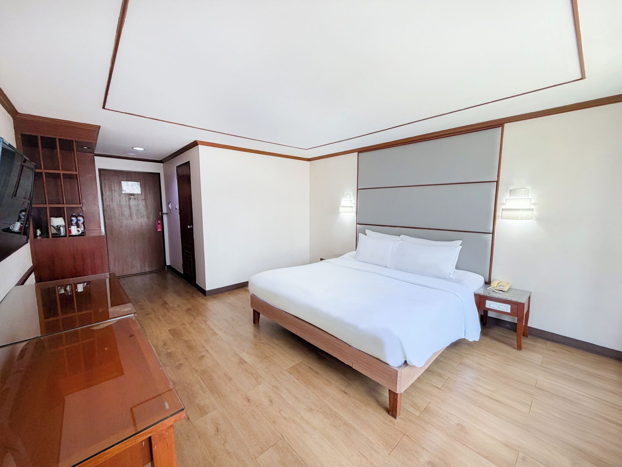 A hotel room with a wooden bed, a bedside table with a lamp, a wooden dresser, and a television.