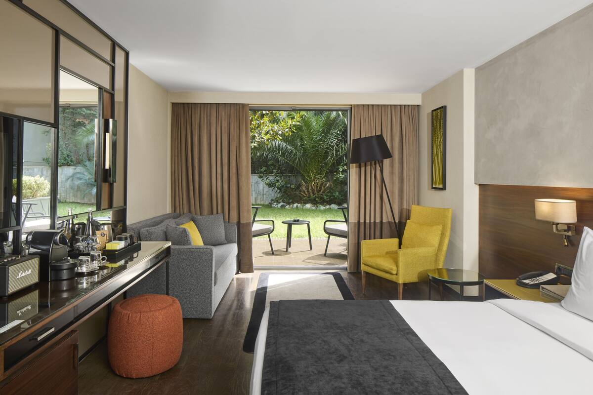 A hotel room with a bed, a desk with a computer, a sofa, a yellow armchair, and a view of the outdoors.