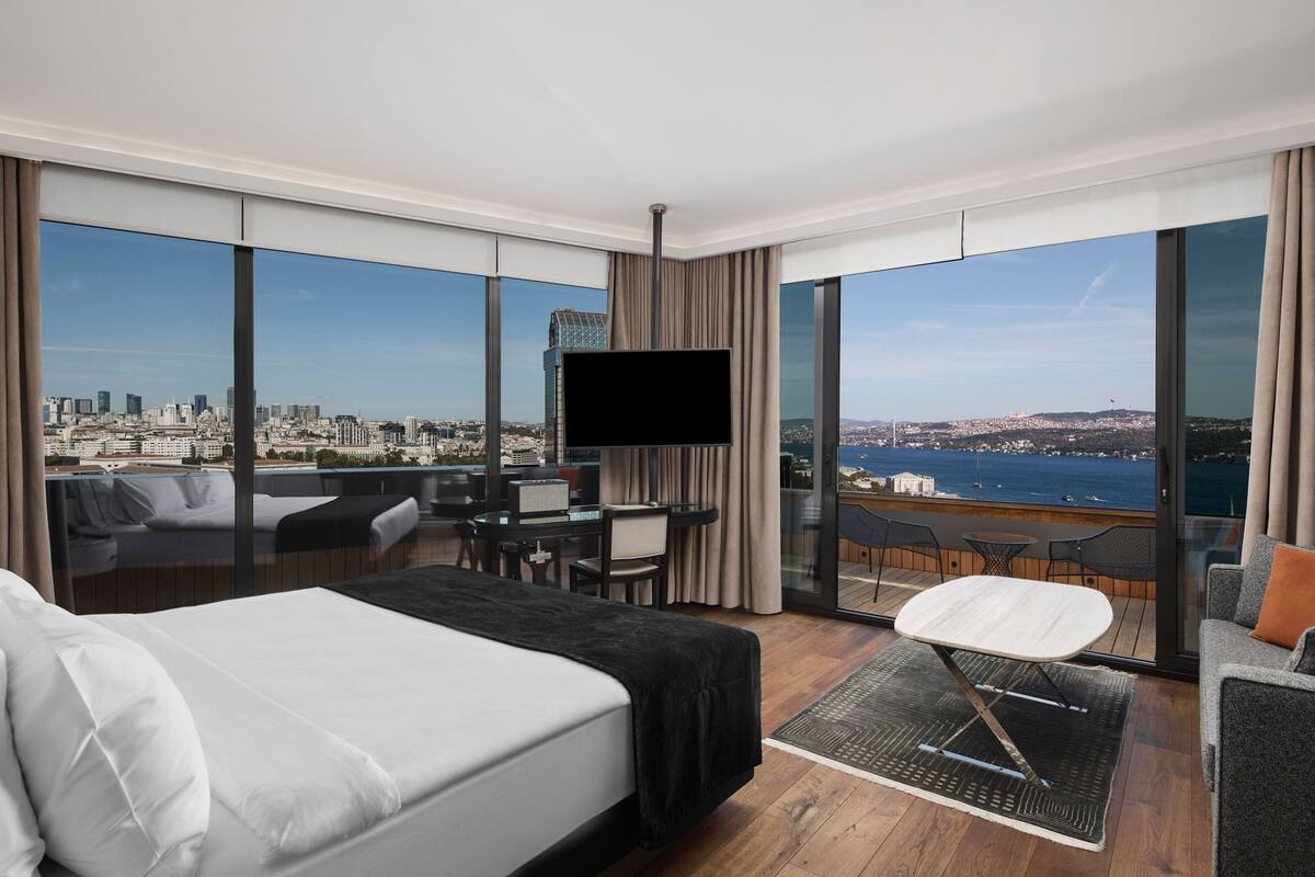 A modern hotel room with a large bed, a desk, and a TV. It features a city view through large windows.