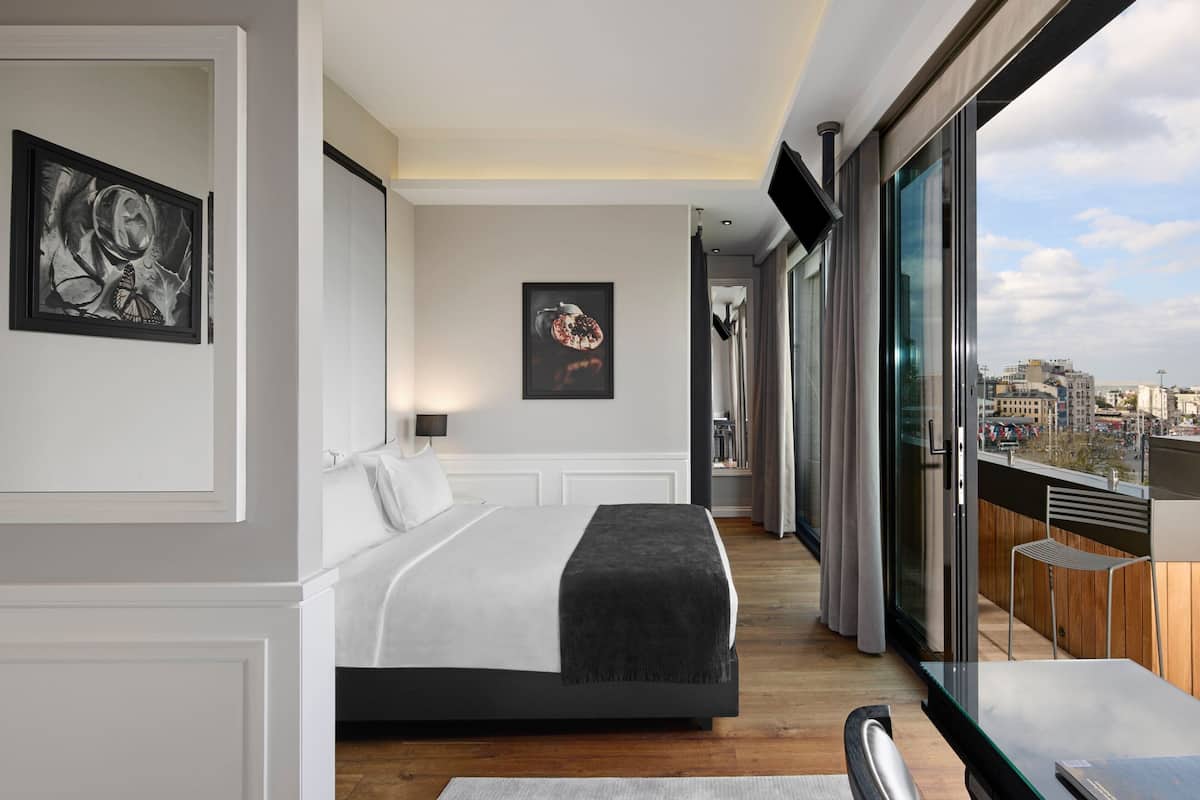 A modern hotel room with a large bed, a balcony with a view of the city, and artwork on the walls.
