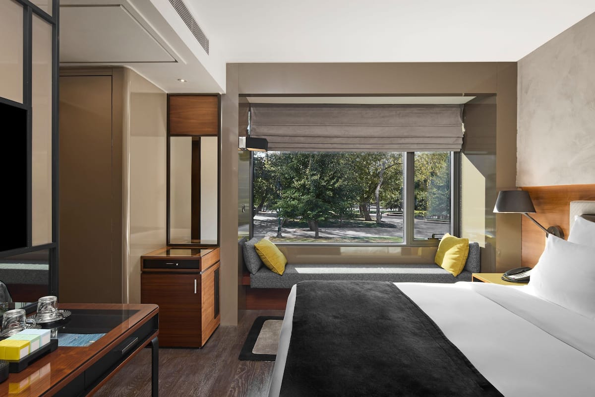 A modern hotel room with a large bed, a desk with a telephone, a seating area with a view of trees, and a window with a view of the outdoors.
