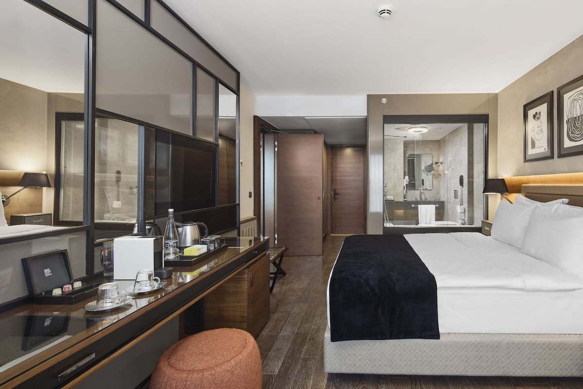 A hotel room with a bed, a desk with a computer and coffee maker, a mirror, and a bathroom visible through a glass door.