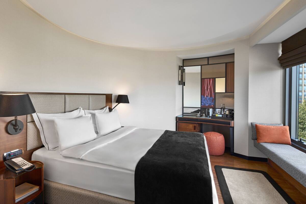 A modern hotel room with a large bed, bedside lamps, a desk with a mirror, and a seating area by the window.