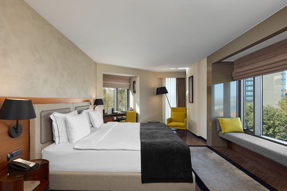 A hotel room with a large bed, a desk with a lamp, a chair, and a window with a view of buildings.