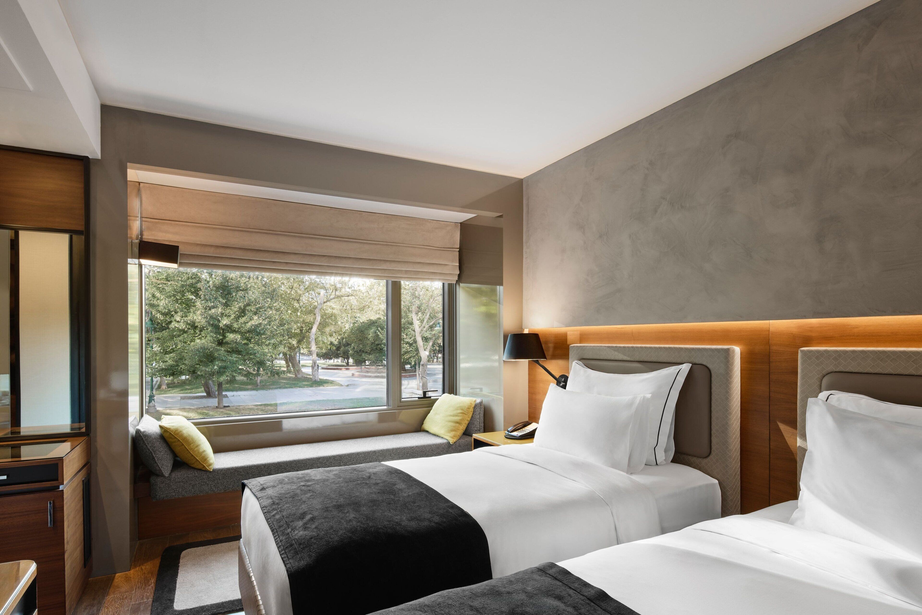 A hotel room with two beds, a large window with a view of trees, a sofa, and a desk with a lamp.