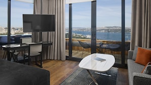 A modern hotel room with a large TV, a desk with a chair, a sofa, and a balcony with a view of the sea and cityscape. - Gezi Hotel Bosphorus, Istanbul, a Member of Design Hotels - Special Class (Istanbul)