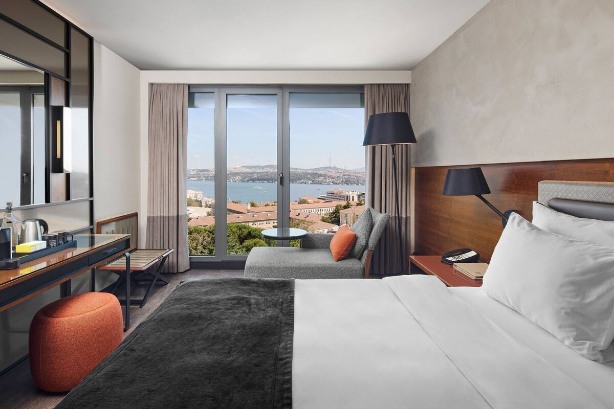 A hotel room with a large bed, a desk with a chair, a small table, and a sofa with a view of the city and water.