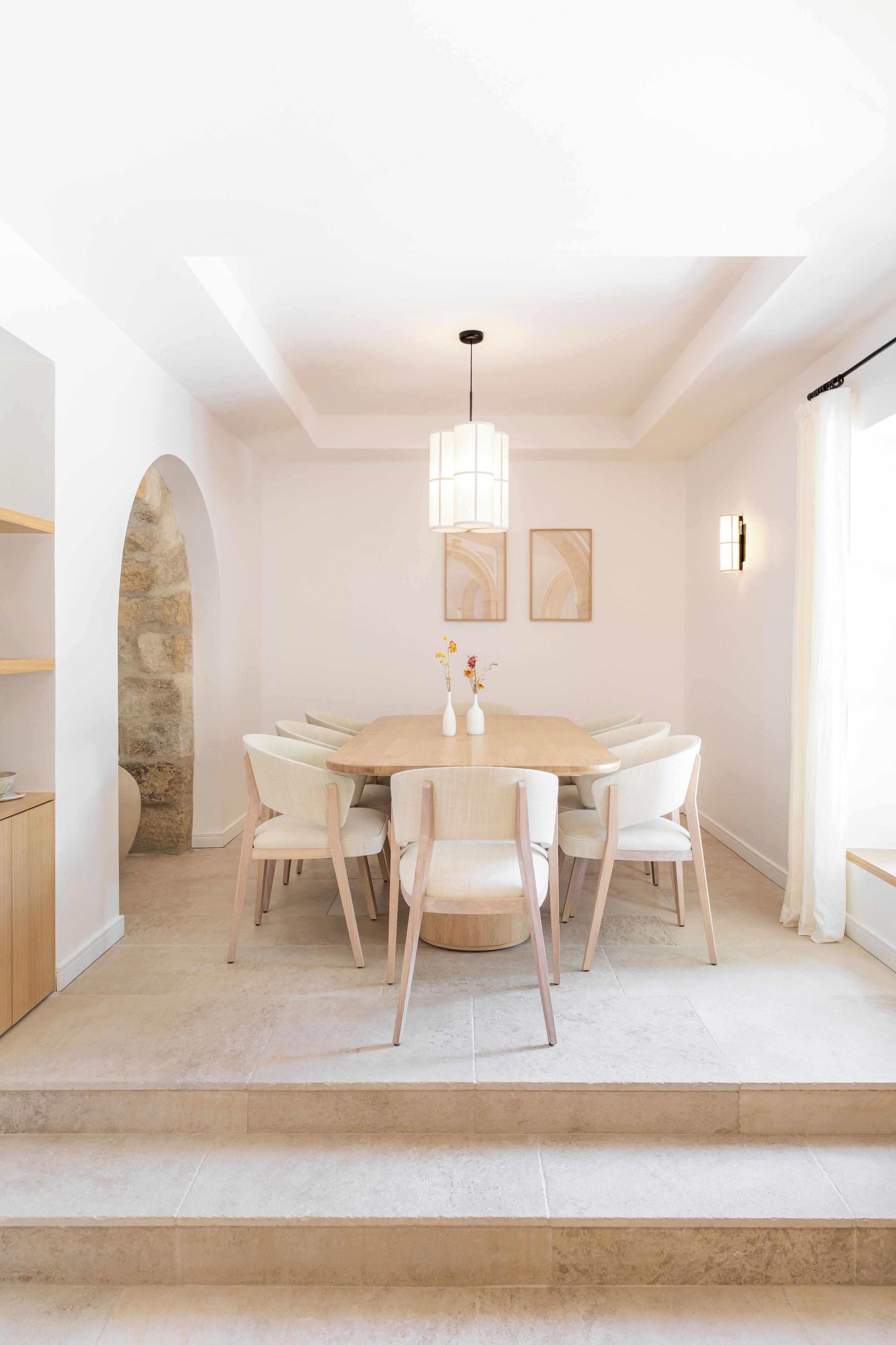Un espace repas comprenant une table ronde, des chaises blanches, un luminaire suspendu et une arche en pierre.