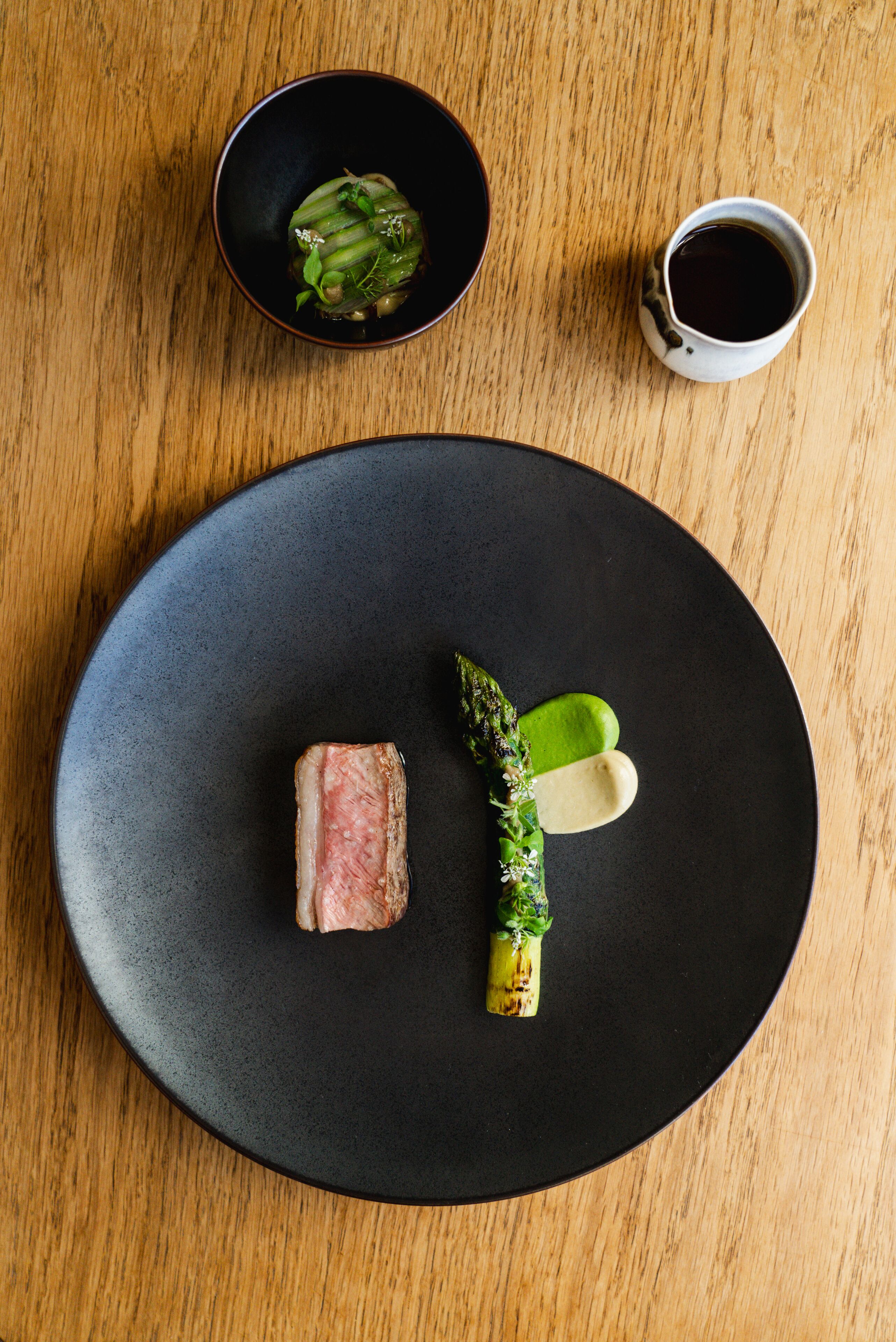 A black plate with a piece of meat, asparagus, and a small bowl of sauce on a wooden table.