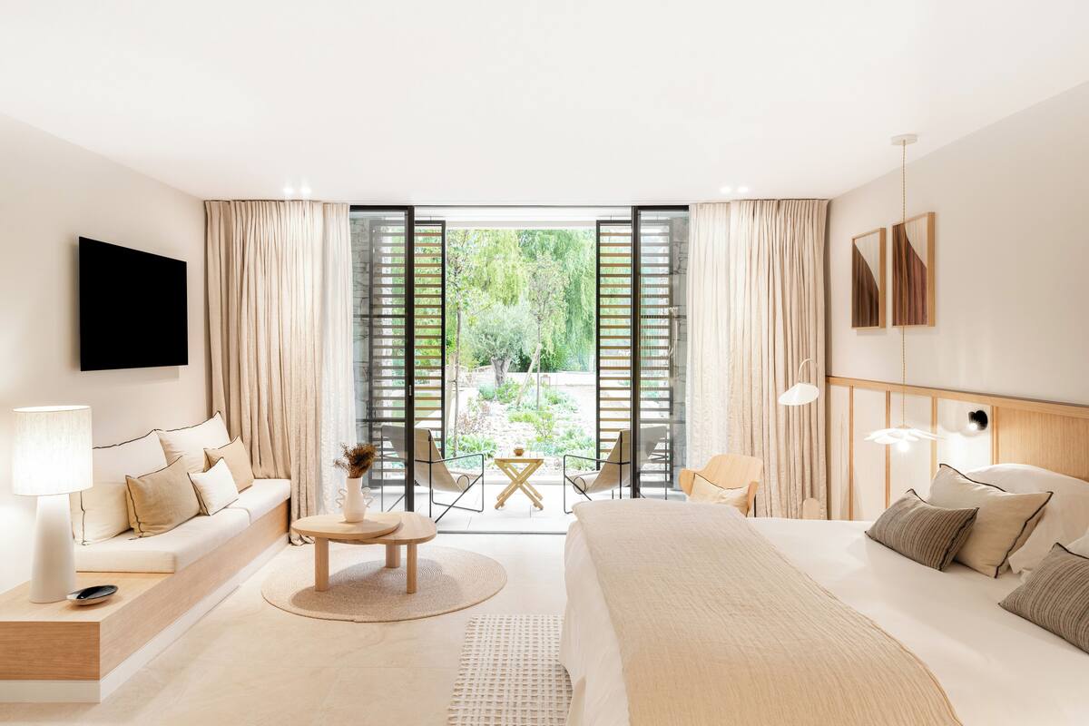 Una habitación de hotel moderna con una cama grande, un sofá, una mesita y un televisor. Incluye un balcón con vistas al verdor.
