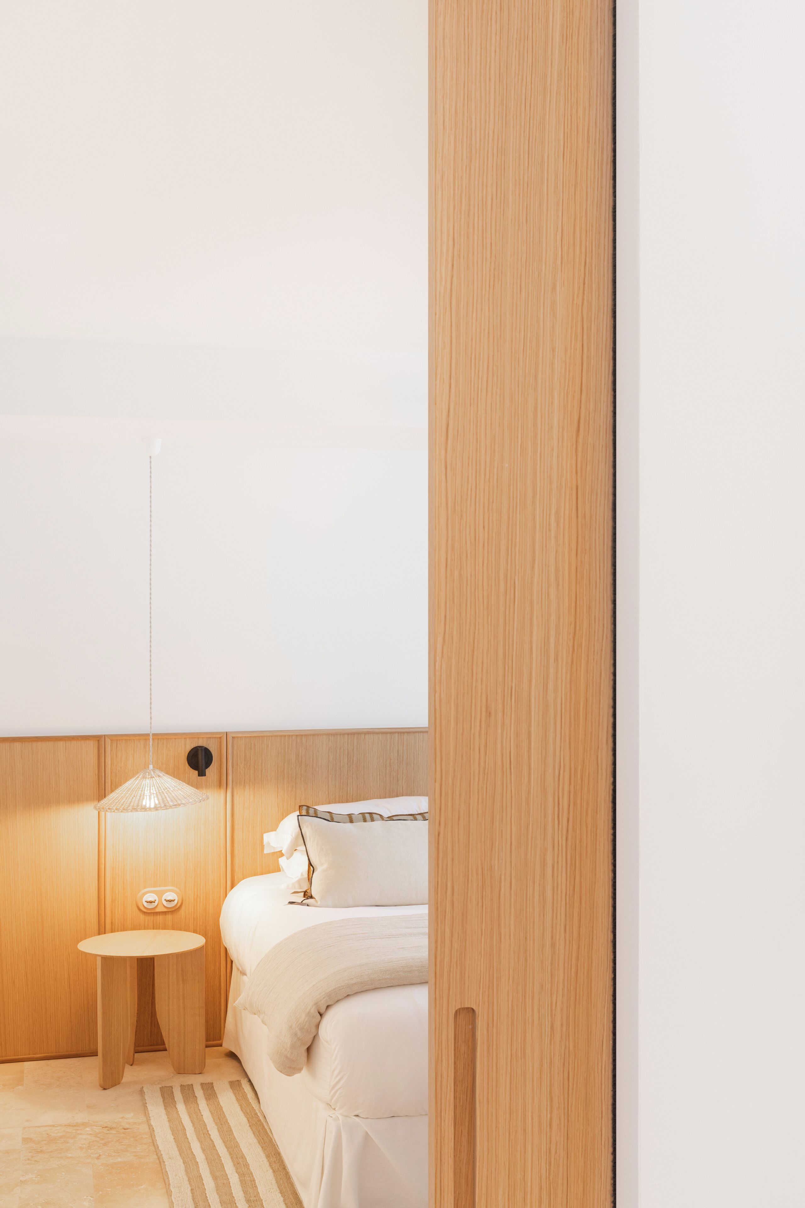 A modern bedroom with a wooden headboard, a bedside table, a lamp, and a bed with white bedding.