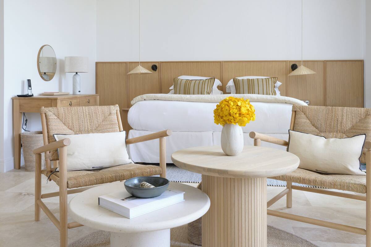 Una habitación de hotel moderna con una cama, dos sillones tapizados, una mesa redonda, una lámpara de noche, un espejo y un jarrón con flores amarillas.
