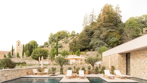 Piscina com espreguiçadeiras e guarda-sóis, cercada por muros de pedra e vegetação.