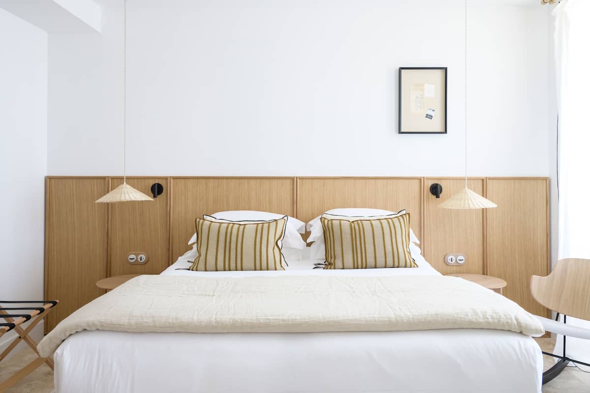 Un dormitorio moderno con cabecera de madera, cama con ropa de cama blanca, almohadas a rayas, dos mesitas de noche y una lámpara colgante.