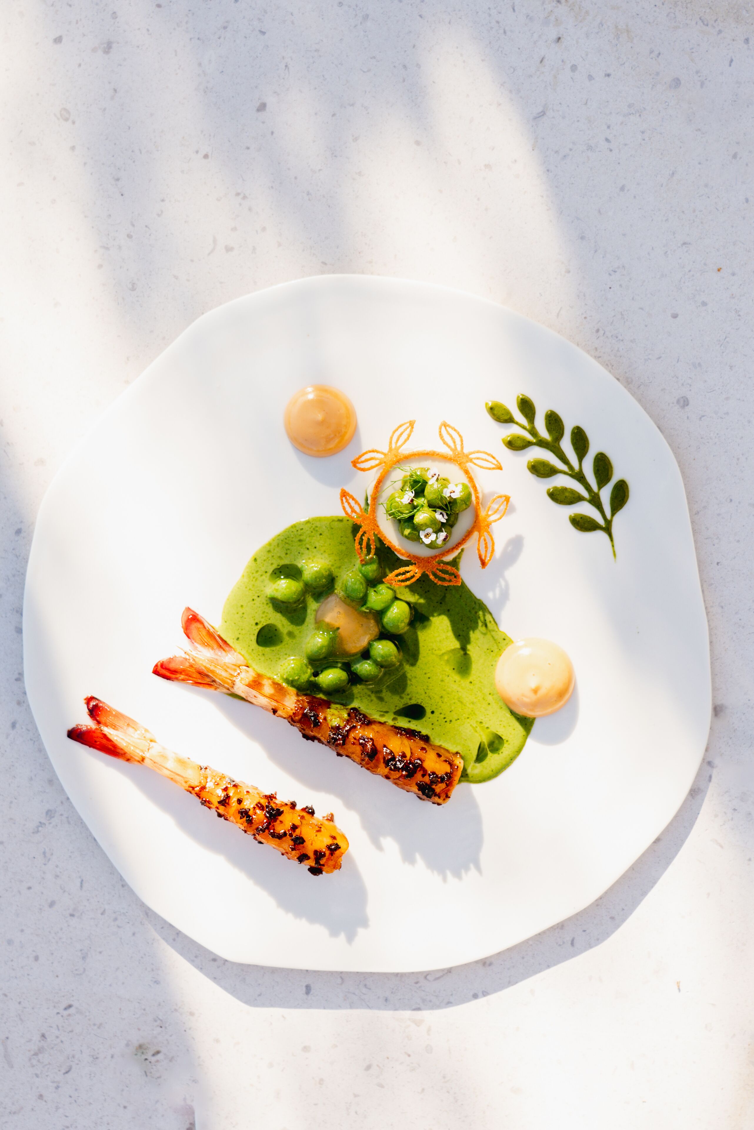 Une assiette contenant un plat de crevettes grillées, de petits pois et une sauce verte, décoré d’une feuille et de filaments d’orange.
