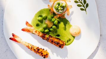 Une assiette contenant un plat de crevettes grillées, de petits pois et une sauce verte, décoré d’une feuille et de filaments d’orange.