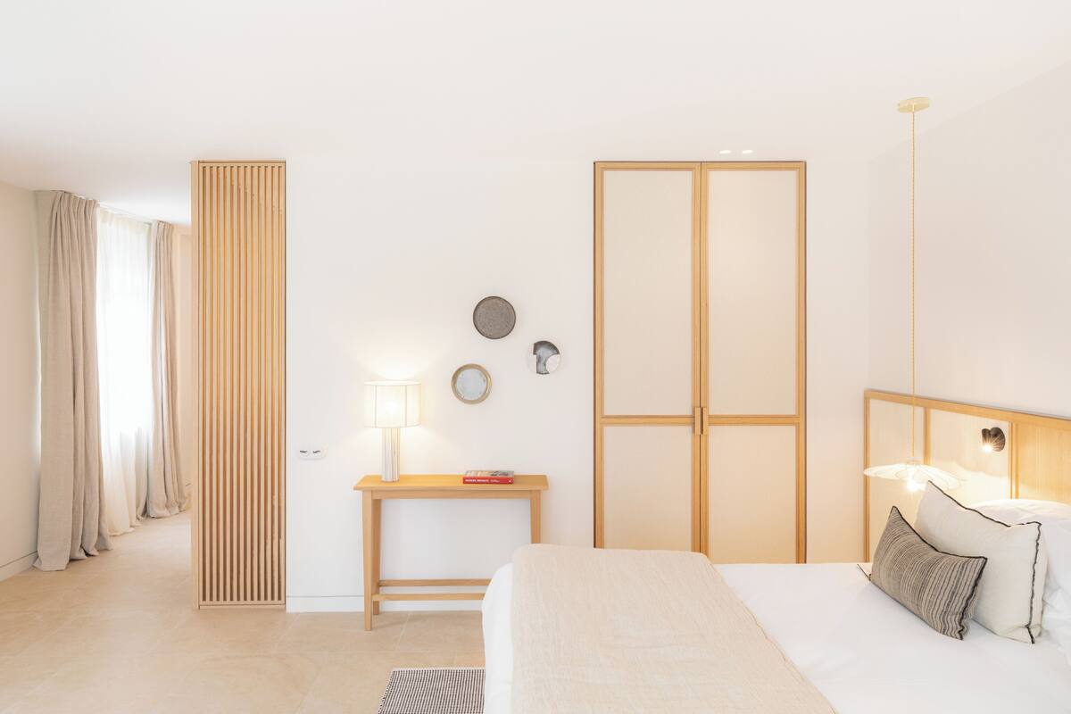 Una habitación de hotel moderna con una cama, mesita de noche, lámpara y vistas al pasillo.