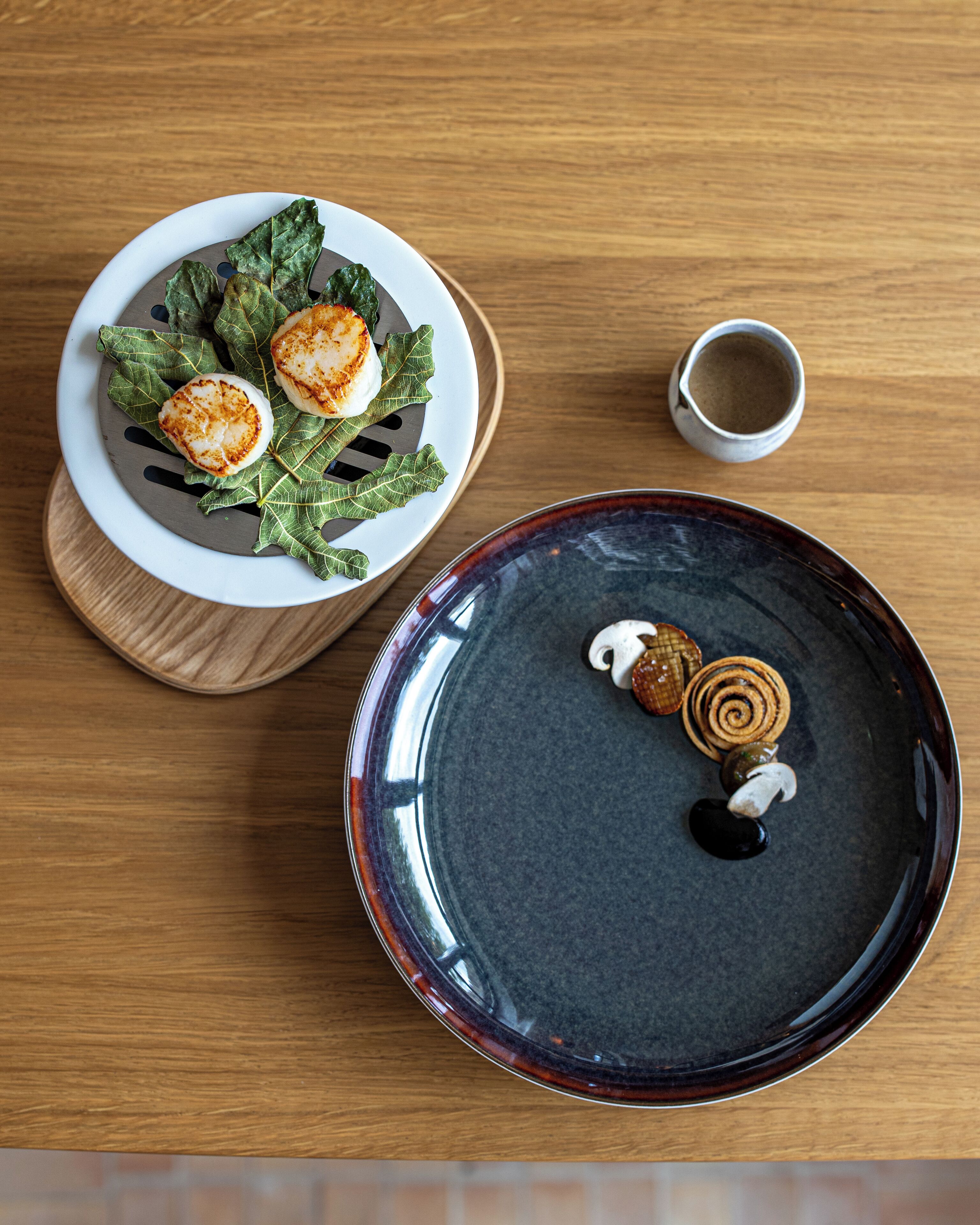 Uma mesa de madeira com dois pratos de comida, um com vieiras e vegetais, e outro com um prato escuro e um item em espiral.