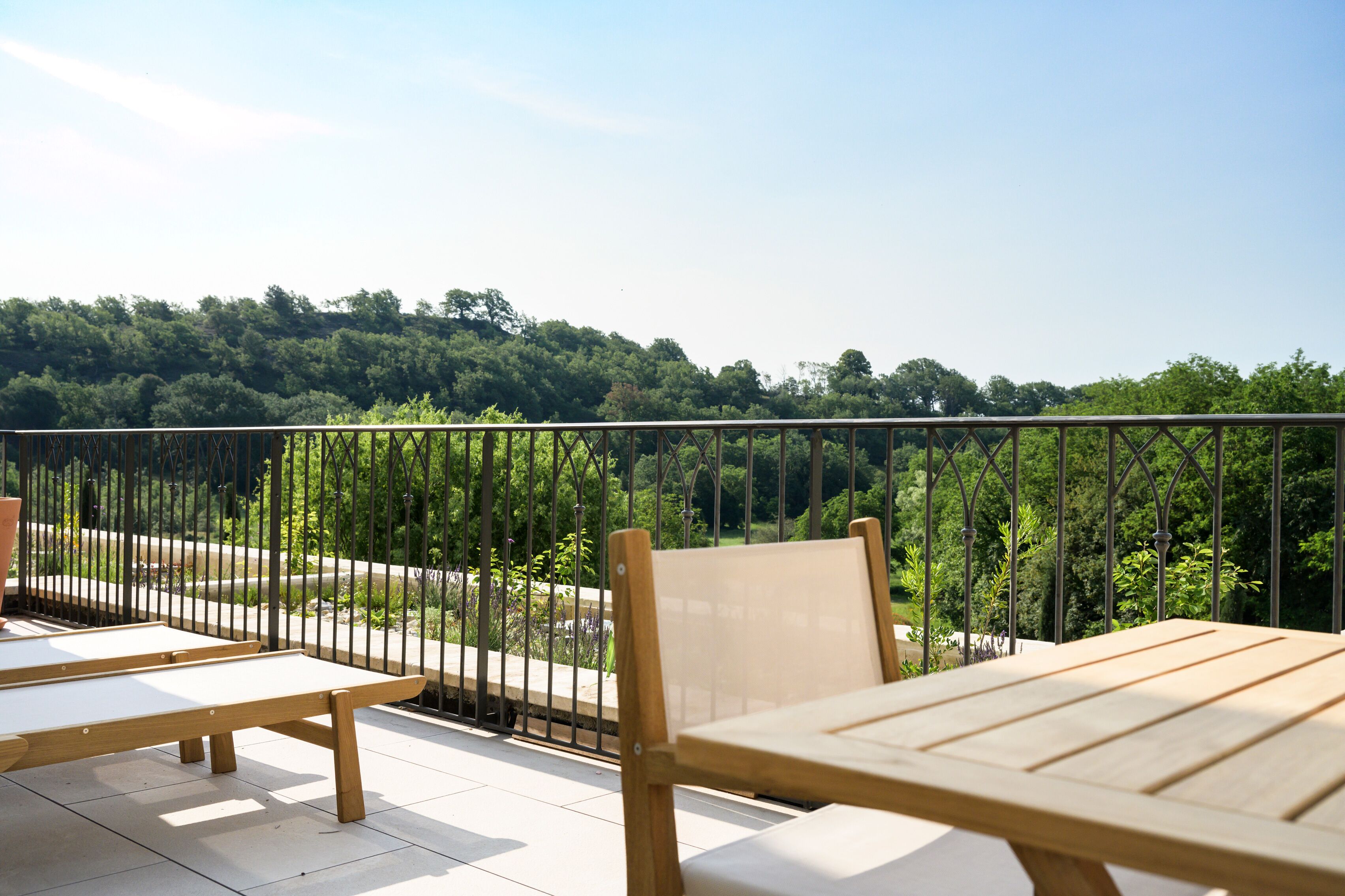 A balcony with wooden furniture and a metal railing overlooking a forested area.