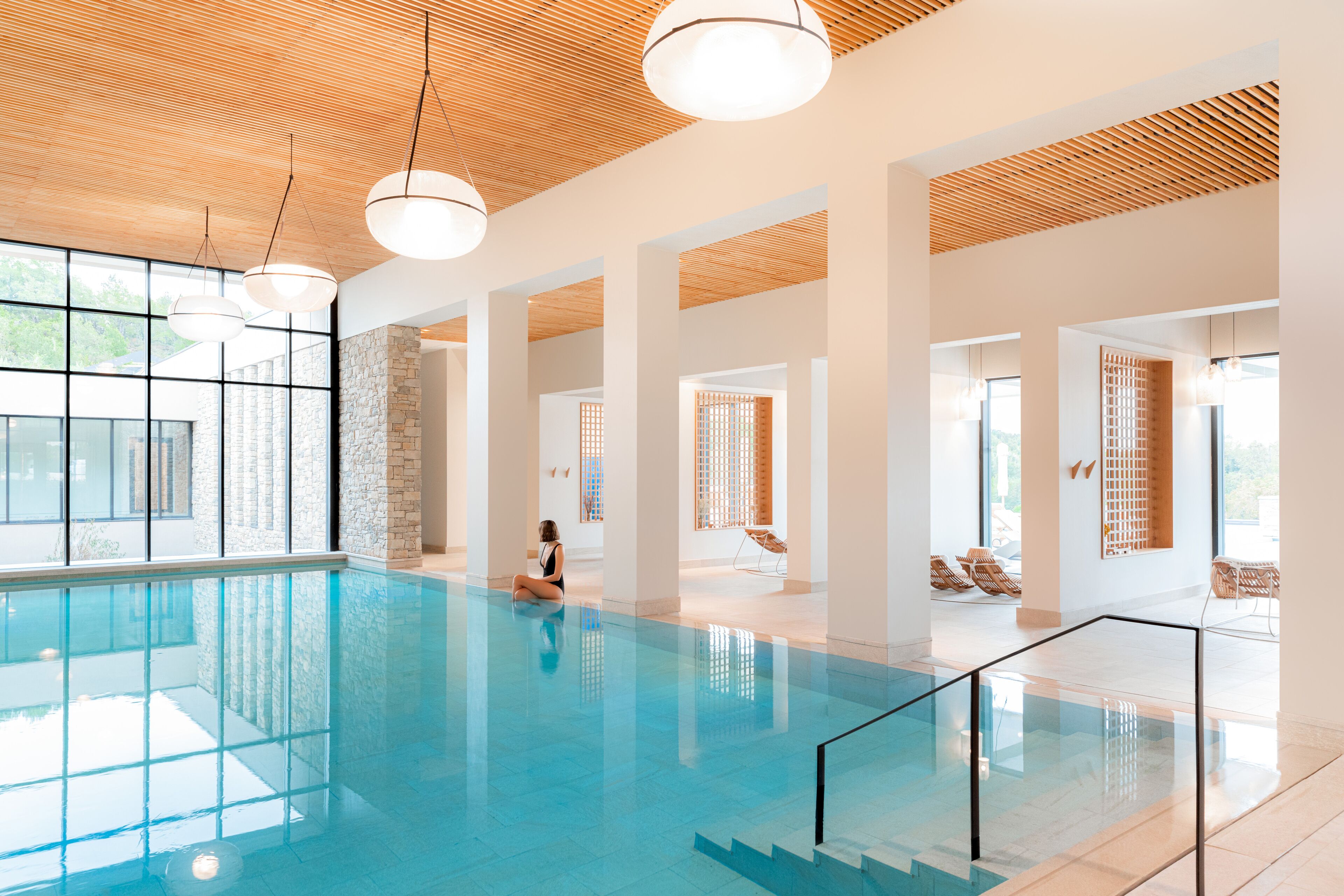 A spacious indoor swimming pool with clear glass walls, wooden ceiling, and modern lighting fixtures.