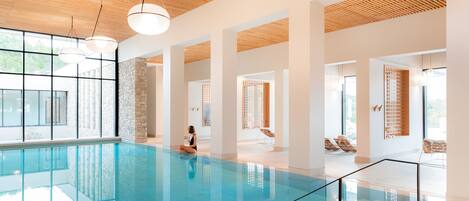 A spacious indoor swimming pool with clear glass walls, wooden ceiling, and modern lighting fixtures.