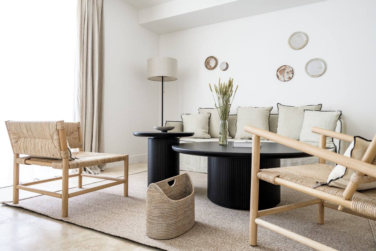 Una sala moderna con una mesa redonda, dos mesitas auxiliares negras, una silla de mimbre, una lámpara de pie y un florero con plantas.