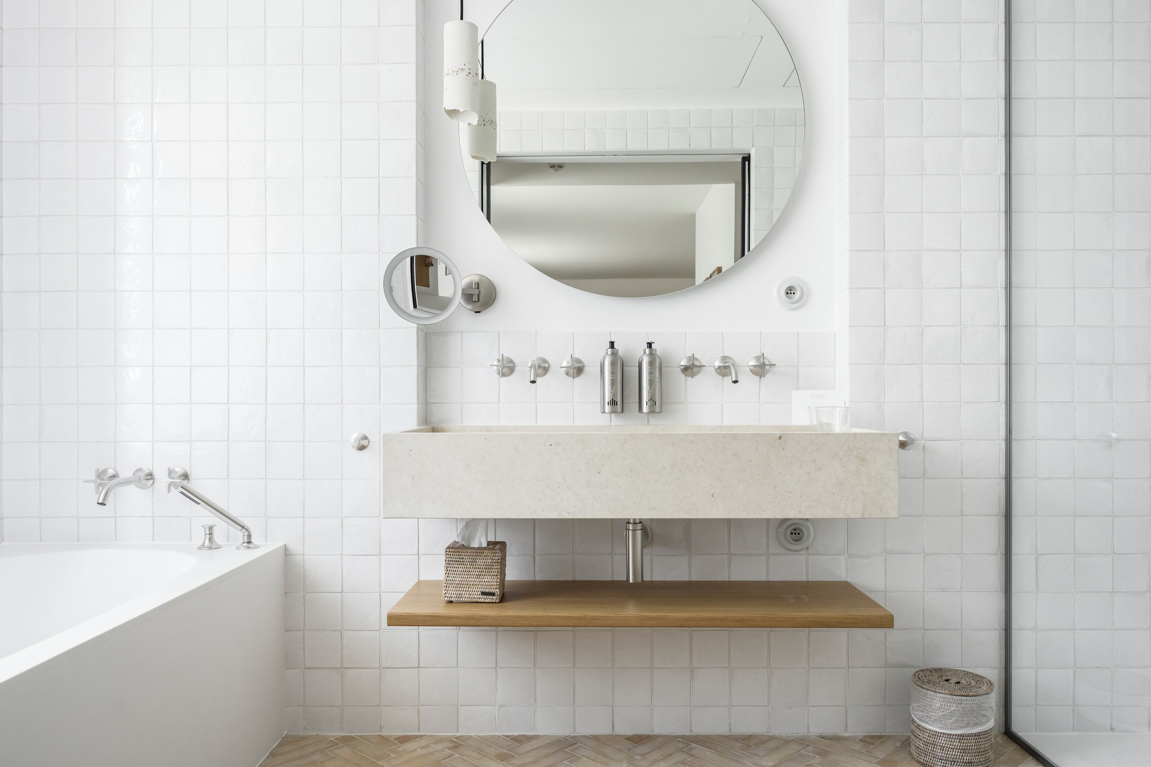 A modern bathroom with a white tiled wall, a round mirror, a white sink with two faucets, and a wooden shelf below the sink.
