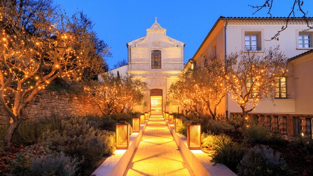 Um caminho leva a uma igreja com torre sineira, cercada por árvores e iluminada por luzes quentes.
