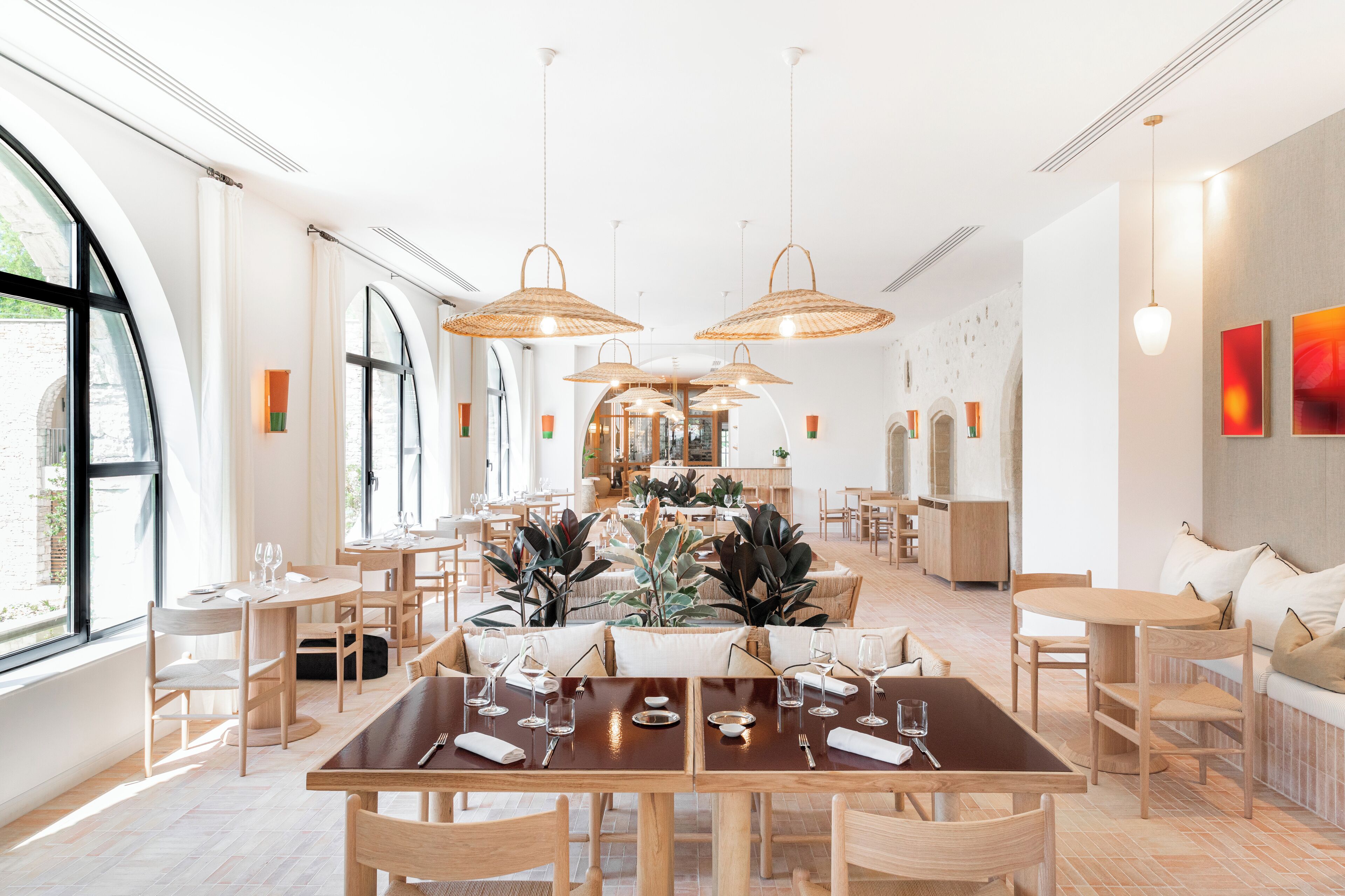 Un comedor amplio con mesas y sillas de madera, ventanas grandes y lámparas de mimbre colgantes.