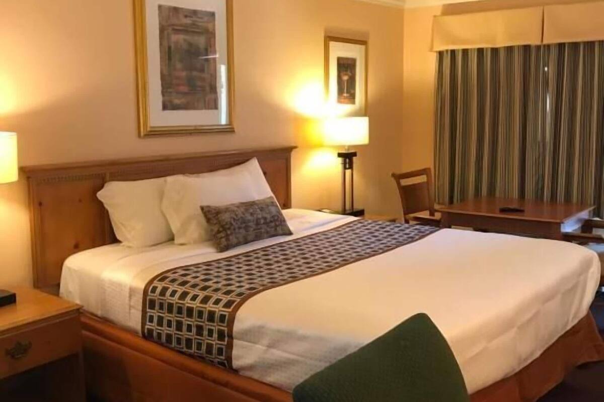 A hotel room with a large bed, two bedside lamps, a framed picture above the bed, a chair, and a curtain.