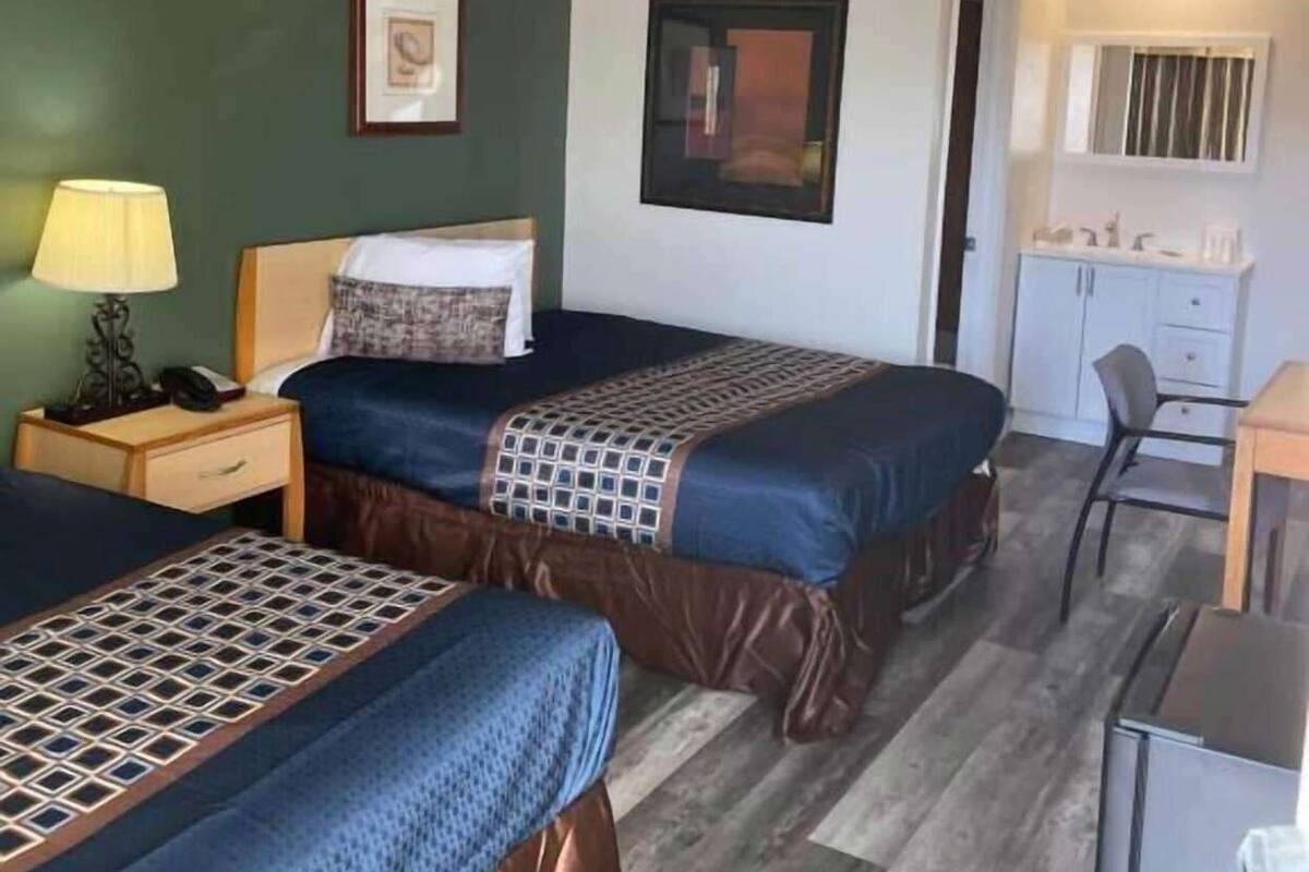 A hotel room with two beds, a desk, a chair, a lamp, and a small kitchenette.