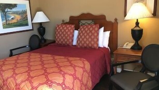A hotel room with a bed, a chair, a nightstand with a lamp, and a painting on the wall. - Sands Motel (Riverside)