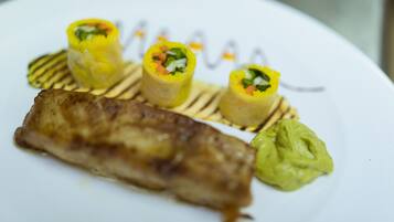 Un plato con un filete de pescado a la plancha, tres rollitos de verdura amarillos y una cucharada de salsa verde.