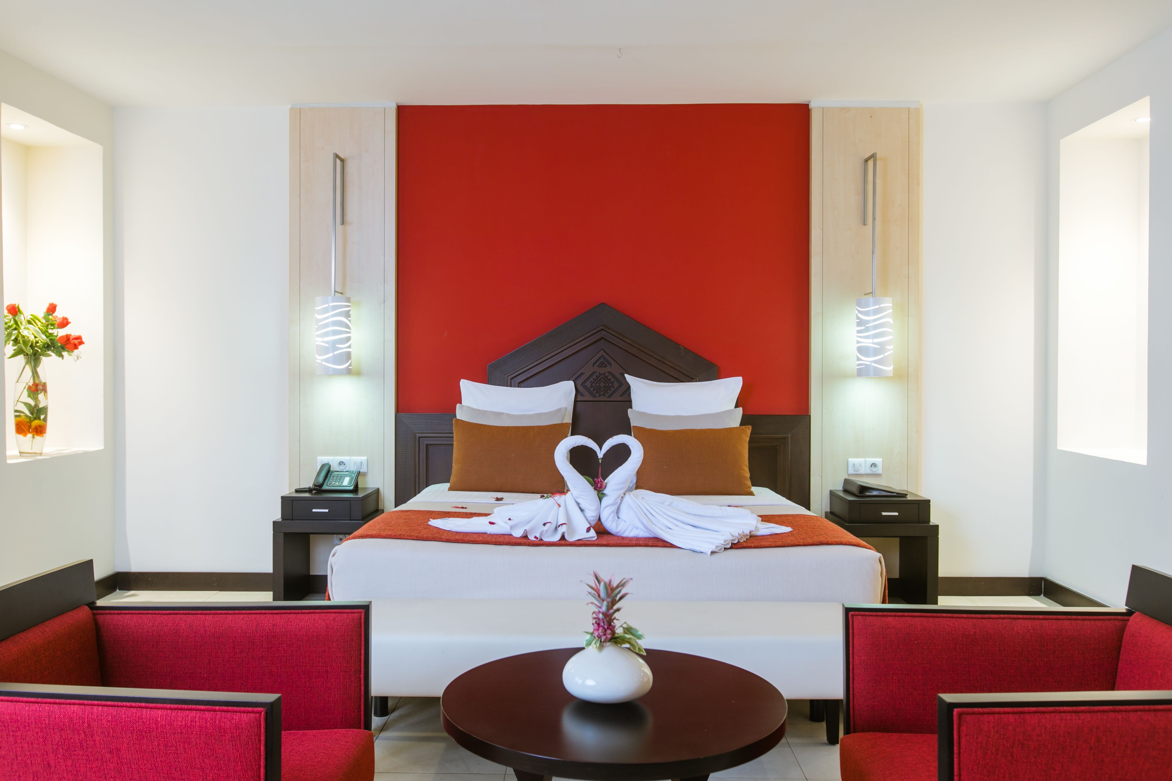 Una habitación de hotel con una cama grande, ropa de cama roja y blanca, una disposición de toallas con forma de corazón, un jarrón con flores y sillones tapizados en rojo.