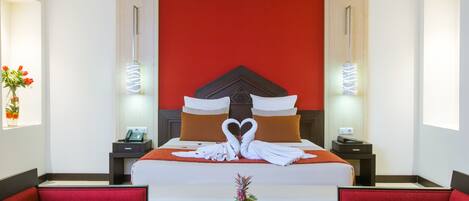Una habitación de hotel con una cama grande, ropa de cama roja y blanca, una disposición de toallas con forma de corazón, un jarrón con flores y sillones tapizados en rojo.