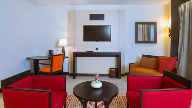 Habitación de hotel con una televisión montada en la pared, una mesita con un florero, dos sillones rojos, un sofá con cojines naranjas y una lámpara sobre una mesita auxiliar.