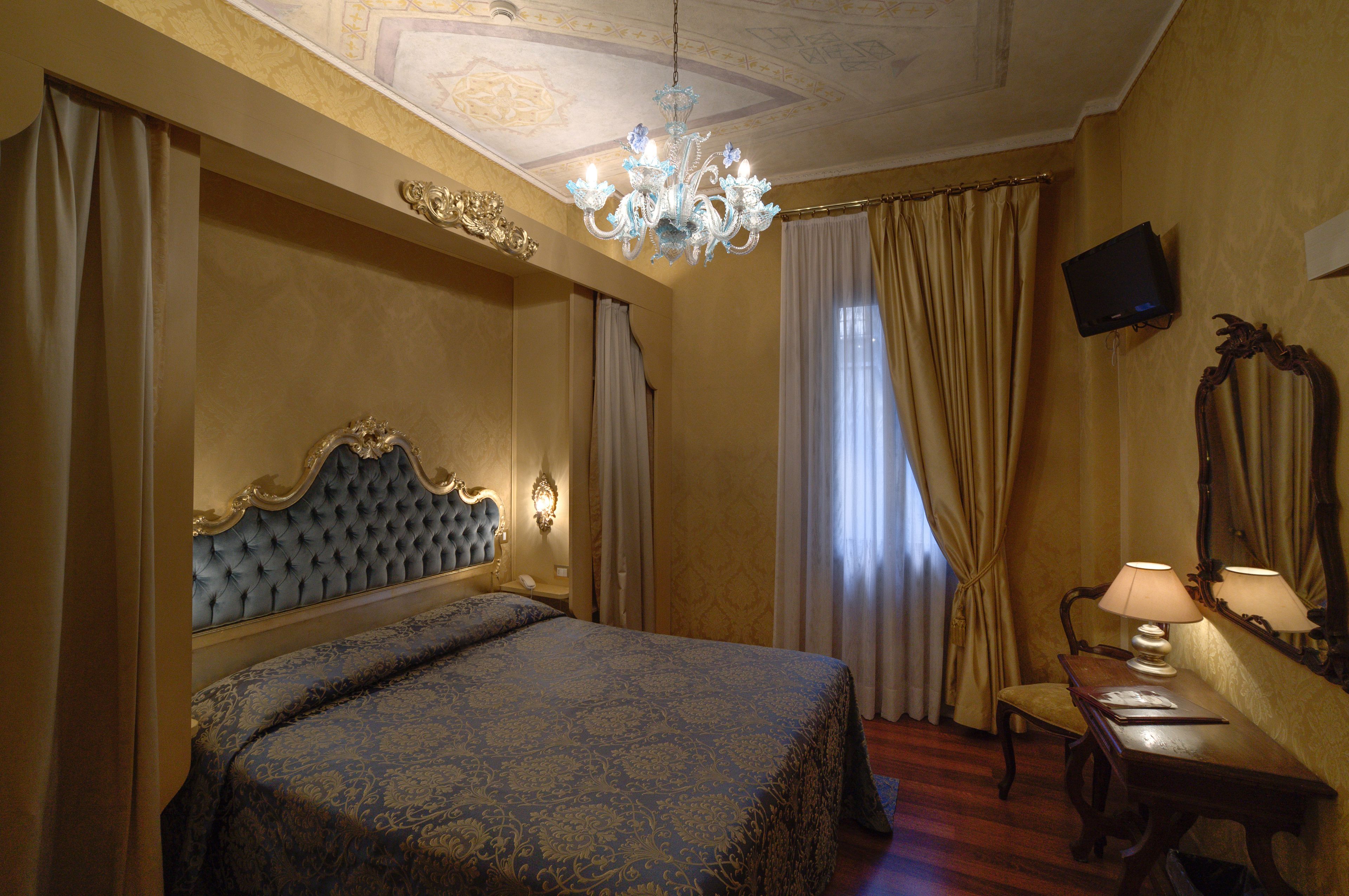 A hotel room with a large bed, a chandelier, a desk with a lamp, and a television on the wall.