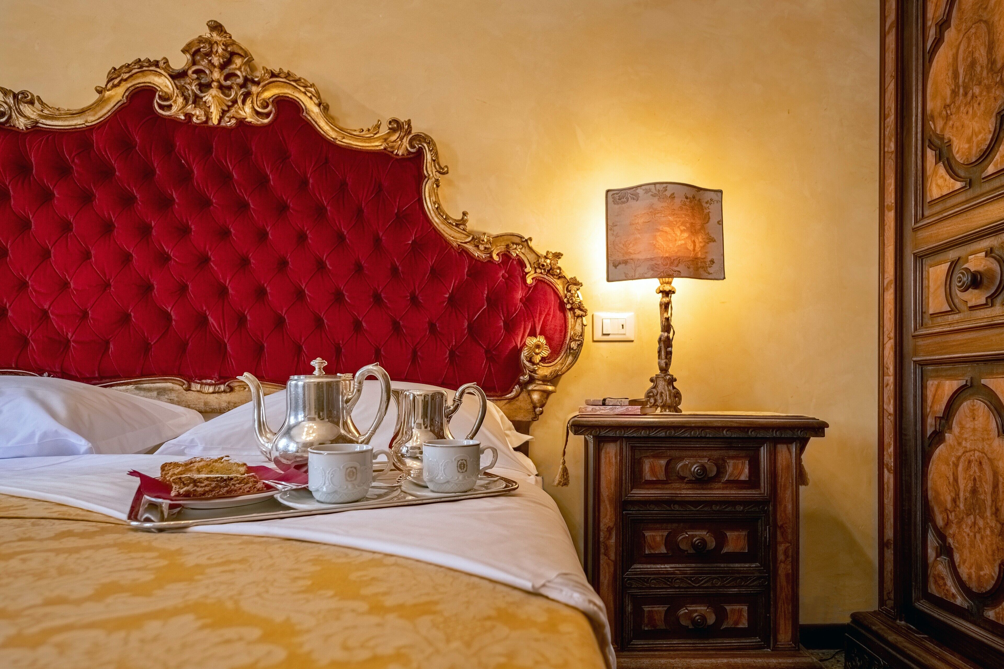 Un'elegante camera da letto con una testiera imbottita rossa, un comodino con una lampada e un vassoio con tazzine e teiera.