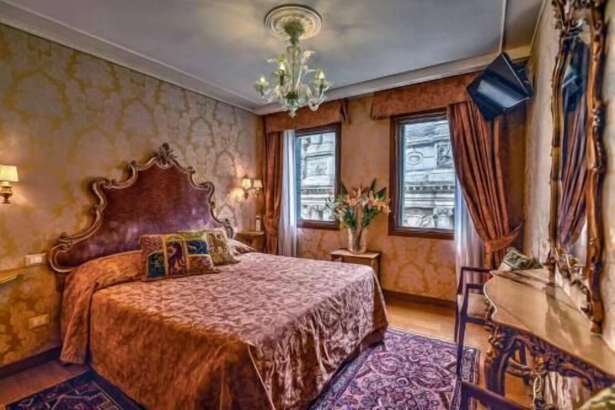 A bedroom with a large bed, a chandelier, a small table with a vase, and a view of buildings through the window.