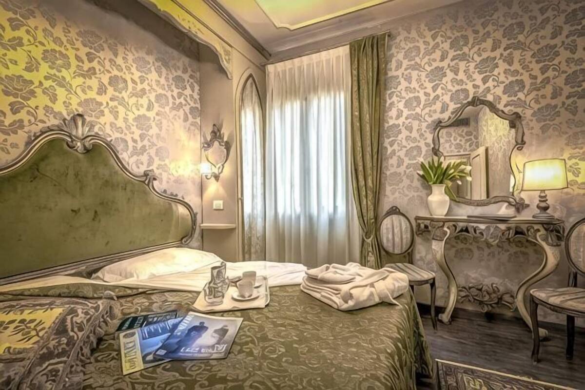 A hotel room with a bed, a bedside table with a lamp, a mirror, a chair, and a window with curtains.