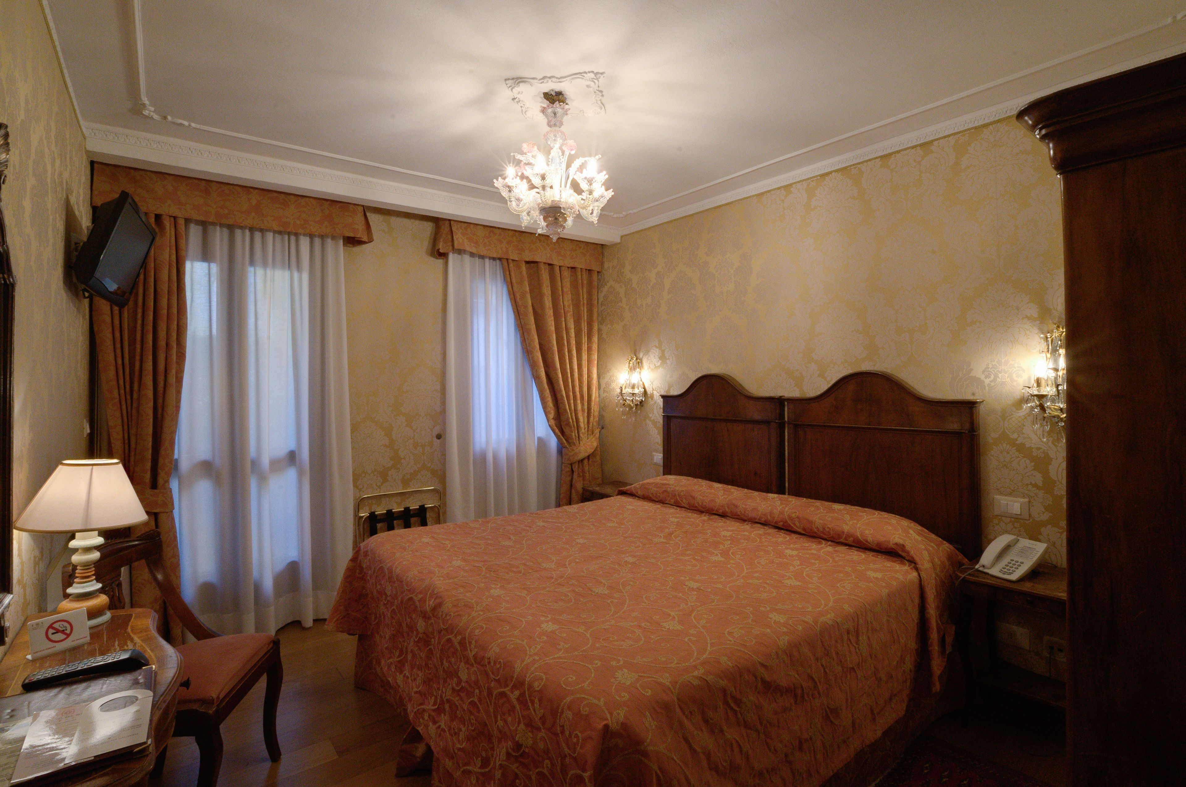 A hotel room with a large bed, a desk with a lamp, a chair, a television mounted on the wall, and a telephone on the bedside table.